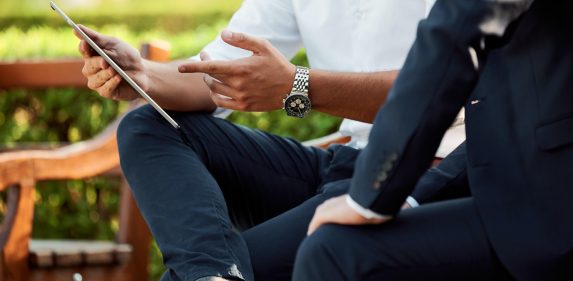 Two people in business attire, one holding a digital tablet, engaged in a discussion outdoors.