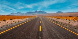 Straight desert road leading towards distant mountains under a clear blue sky.